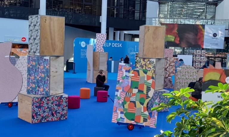Vibrant exhibition hall with colorful abstract panels and wooden structures on a blue floor. People are seated nearby. Bright, modern atmosphere.