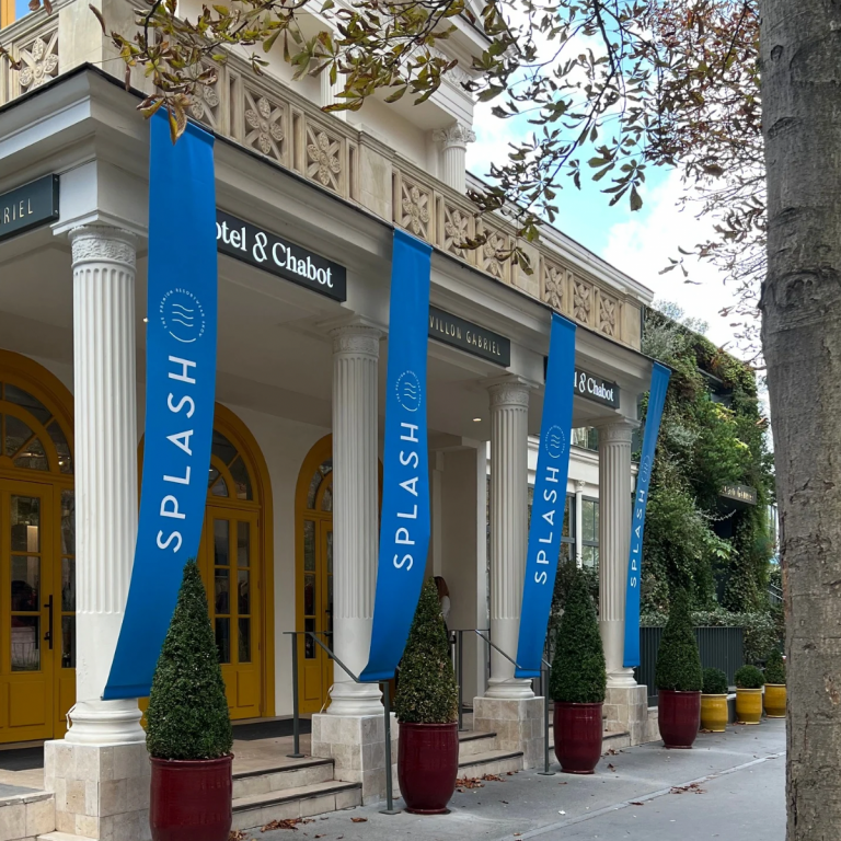 Ornate building entrance trade show premium resort wear with four white columns, blue banners reading "SPLASH," and potted greenery. Bright yellow doors add a cheerful touch.