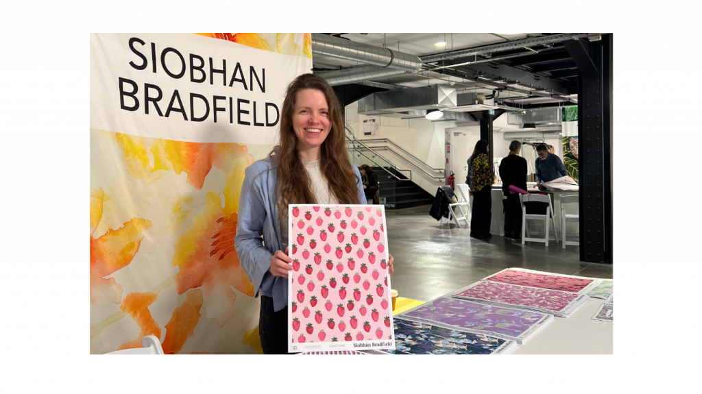 A cheerful woman, Siobhan Bradfield stands at a booth holding a pink patterned poster that is a textile design print with strawberries on paper. Behind her is a banner and table with colorful designs. Industrial interior.