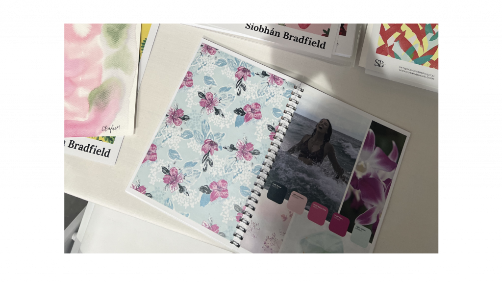 An open spiral notebook displays a floral textile design on a pastel blue background, with swatches and a photo of a woman in waves. Nearby are vibrant fabric samples.