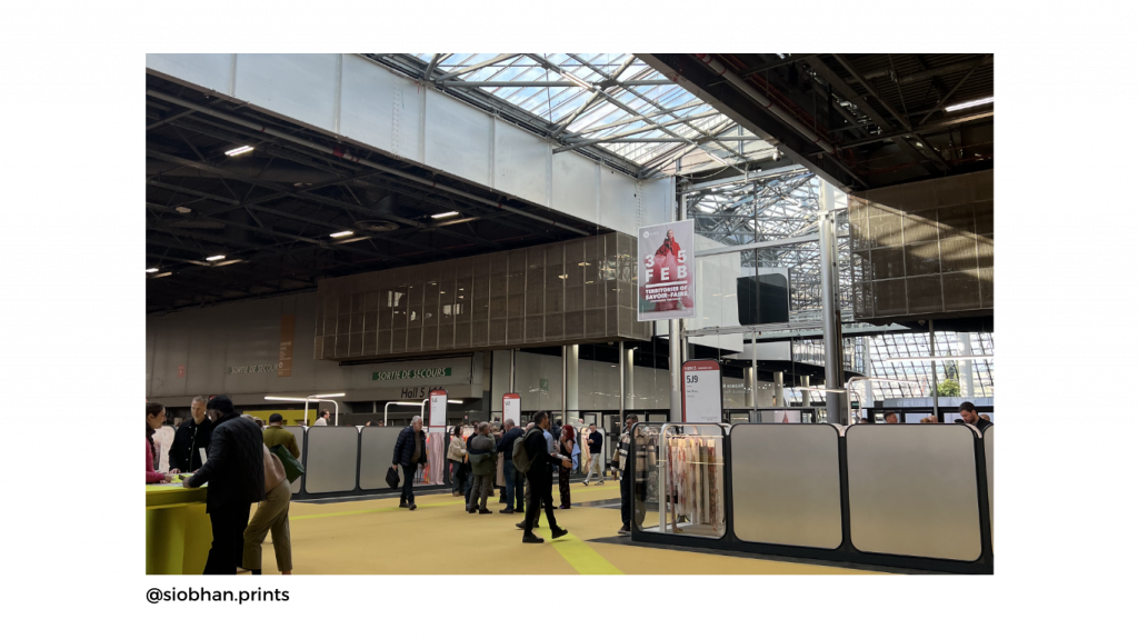 Spacious exhibition hall with people walking and interacting at booths at Premiere Vision a textile and fashion and sourcing design fair. Large skylights provide natural lighting. A sign with "FEB" hangs overhead.